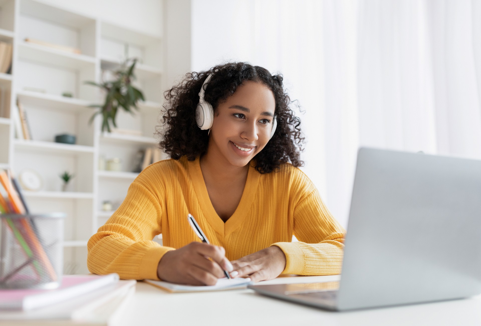 Happy African American woman in headphones having online meeting on laptop, writing down info during distance lesson
